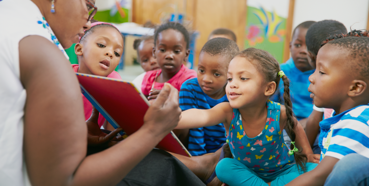 African American children sitting on the ground while listening to a teacher reading them a book